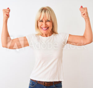 Smiling woman with blonde hair in a white t-shirt flexes her arms and shows her biceps, standing against a plain white background, radiating confidence—perfectly embodying the strength supported by Hormone Replacement experts.