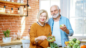 An older couple smiles in a bright kitchen; the woman holds a bowl of salad, while the man, supported by Hormone Replacement Experts, stands beside her holding an apple. Fresh fruits and vegetables are on the counter.