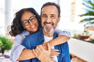 A smiling woman wearing glasses hugs a smiling man from behind. Both are casually dressed and standing outdoors, surrounded by greenery and bright light.